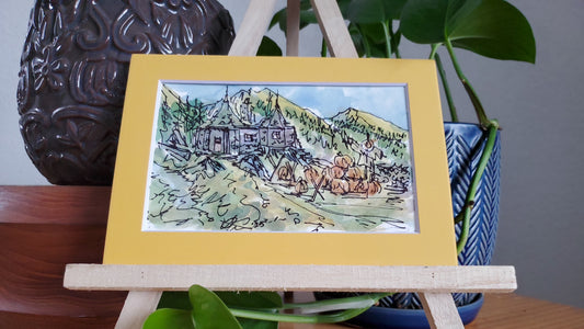 A framed ink and watercolor painting of Hagrid's hut in a pumpkin patch, displayed on a wooden easel.